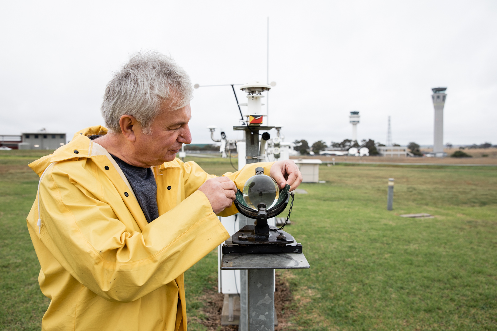 Weather observer in yellow raincoat checks outdoor observation equipment at an airport. 